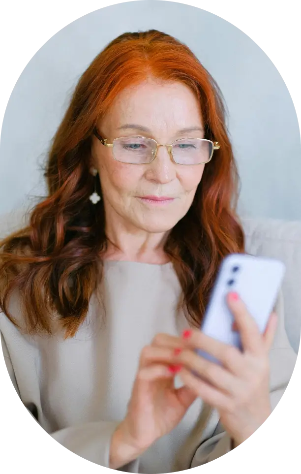 Senior woman with red hair and glasses using a smartphone, representing online appointment booking or patient engagement.